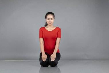 Kırmızı tişörtlü, siyah taytlı, yoga ve esneme egzersizleri yapan sağlıklı genç bir kadın gri arka planda izole edilmiş.