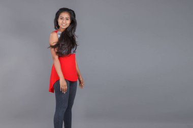 Genç, güzel bir Hintli kadın modelin gündelik kıyafetleriyle stüdyo fotoğrafı. Kırmızı ve gri tasarımcı bluzu ve beyaz arka planda siyah kot pantolon giyiyor. Kadın manken. Moda Portresi.