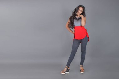 Genç, güzel bir Hintli kadın modelin gündelik kıyafetleriyle stüdyo fotoğrafı. Kırmızı ve gri tasarımcı bluzu ve beyaz arka planda siyah kot pantolon giyiyor. Kadın manken. Moda Portresi.