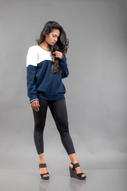 Gri arka planda günlük kışlık giysili Hintli genç bayan model. Siyah tayt giyen uzun siyah saçlı bir manken, mavi beyaz bir kazak..