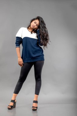 Gri arka planda günlük kışlık giysili Hintli genç bayan model. Siyah tayt giyen uzun siyah saçlı bir manken, mavi beyaz bir kazak..