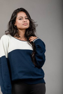 Gri arka planda günlük kışlık giysili Hintli genç bayan model. Siyah tayt giyen uzun siyah saçlı bir manken, mavi beyaz bir kazak..