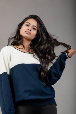 Gri arka planda günlük kışlık giysili Hintli genç bayan model. Siyah tayt giyen uzun siyah saçlı bir manken, mavi beyaz bir kazak..