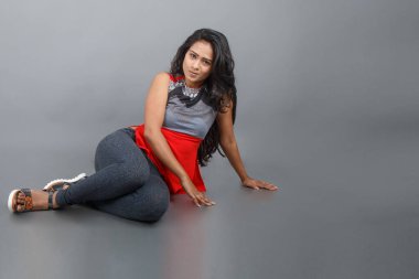 Gri arka planda kırmızı ve gri renkli ve mavi kot pantolon giyen, gündelik giyimli genç ve güzel bir Hintli kadın modelin stüdyo fotoğrafı. Kadın manken. Moda Portresi.