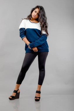 Gri arka planda günlük kışlık giysili Hintli genç bayan model. Siyah tayt giyen uzun siyah saçlı bir manken, mavi beyaz bir kazak..