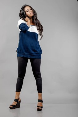 Gri arka planda günlük kışlık giysili Hintli genç bayan model. Siyah tayt giyen uzun siyah saçlı bir manken, mavi beyaz bir kazak..