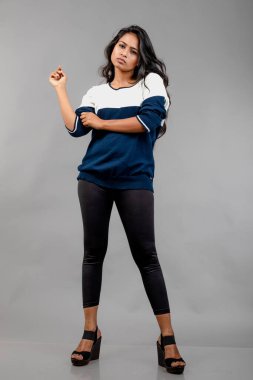 Gri arka planda günlük kışlık giysili Hintli genç bayan model. Siyah tayt giyen uzun siyah saçlı bir manken, mavi beyaz bir kazak..