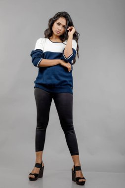 Gri arka planda günlük kışlık giysili Hintli genç bayan model. Siyah tayt giyen uzun siyah saçlı bir manken, mavi beyaz bir kazak..