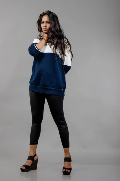 Indian young female model in casual winterwear against grey background. Long black haired model wearing black leggings, blue and white sweatshirt.