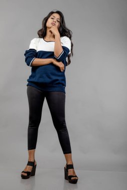 Gri arka planda günlük kışlık giysili Hintli genç bayan model. Siyah tayt giyen uzun siyah saçlı bir manken, mavi beyaz bir kazak..