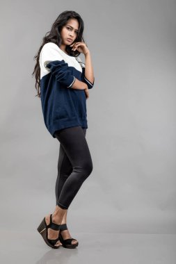 Gri arka planda günlük kışlık giysili Hintli genç bayan model. Siyah tayt giyen uzun siyah saçlı bir manken, mavi beyaz bir kazak..