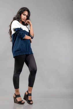 Gri arka planda günlük kışlık giysili Hintli genç bayan model. Siyah tayt giyen uzun siyah saçlı bir manken, mavi beyaz bir kazak..