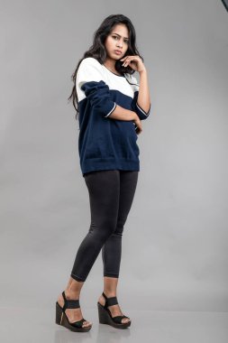 Gri arka planda günlük kışlık giysili Hintli genç bayan model. Siyah tayt giyen uzun siyah saçlı bir manken, mavi beyaz bir kazak..