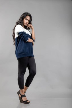 Gri arka planda günlük kışlık giysili Hintli genç bayan model. Siyah tayt giyen uzun siyah saçlı bir manken, mavi beyaz bir kazak..