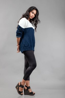 Gri arka planda günlük kışlık giysili Hintli genç bayan model. Siyah tayt giyen uzun siyah saçlı bir manken, mavi beyaz bir kazak..