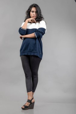 Gri arka planda günlük kışlık giysili Hintli genç bayan model. Siyah tayt giyen uzun siyah saçlı bir manken, mavi beyaz bir kazak..