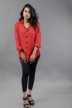 Gri arka plan karşısında gündelik giyimli Hintli genç bayan model - stok fotoğrafı. Kırmızı gömlek ve siyah kot giyen uzun siyah saçlı bir manken..