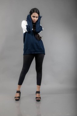 Gri arka planda günlük kışlık giysili Hintli genç bayan model. Siyah tayt giyen uzun siyah saçlı bir manken, mavi beyaz bir kazak..