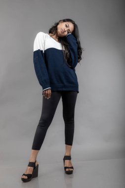 Gri arka planda günlük kışlık giysili Hintli genç bayan model. Siyah tayt giyen uzun siyah saçlı bir manken, mavi beyaz bir kazak..