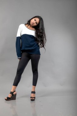 Gri arka planda günlük kışlık giysili Hintli genç bayan model. Siyah tayt giyen uzun siyah saçlı bir manken, mavi beyaz bir kazak..