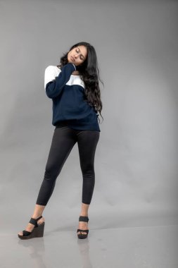 Gri arka planda günlük kışlık giysili Hintli genç bayan model. Siyah tayt giyen uzun siyah saçlı bir manken, mavi beyaz bir kazak..