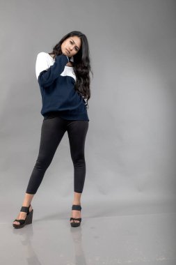 Gri arka planda günlük kışlık giysili Hintli genç bayan model. Siyah tayt giyen uzun siyah saçlı bir manken, mavi beyaz bir kazak..