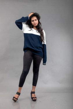 Gri arka planda günlük kışlık giysili Hintli genç bayan model. Siyah tayt giyen uzun siyah saçlı bir manken, mavi beyaz bir kazak..