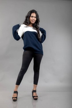 Gri arka planda günlük kışlık giysili Hintli genç bayan model. Siyah tayt giyen uzun siyah saçlı bir manken, mavi beyaz bir kazak..