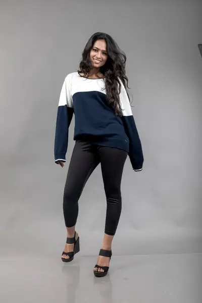 Indian young female model in casual winterwear against grey background. Long black haired model wearing black leggings, blue and white sweatshirt.