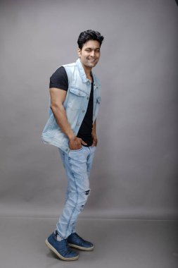 Kısa siyah saçlı, siyah tişörtlü, mavi kot ceketli ve mavi yırtık kot pantolonlu gri arka planda poz veren Hintli genç bir manken. Moda Portresi, Reklam Çekimi.
