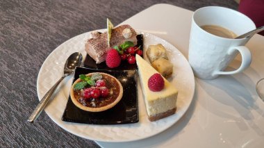 A plate of assorted desserts, including a chocolate tart with berries, a slice of cheesecake with a raspberry, a piece of chocolate with sprinkles, and a small pastry, accompanied by a cup of coffee on a textured white table. clipart