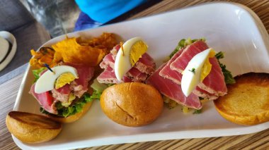 Kurutulmuş ton balığı, marul, bir dilim haşlanmış yumurta ve beyaz dikdörtgen bir tabakta gevrek altın-kahverengi cipslerle servis edilen bir dal bitkiden oluşan üç küçük sandviçten oluşan bir gurme yemeği..