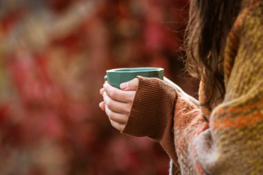 Yeşil kupa tutan eller sonbahar çayı havası kırmızı oliaj bulanık arka plan açık havada Sabah içkisi rahat konsept, arka planda tatil battaniyesi