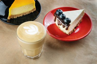 Çikolata kaplı pasta, yaban mersiniyle süslenmiş, el işi kağıdında kurabiyeler. Yanında bir fincan kahve var. Arka planda da bir kek daha var..