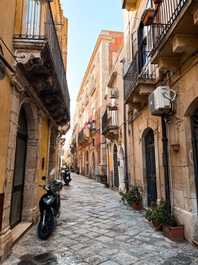 Narrow streets on the old town of Syracuse in Sicily, Italy. Gorgeous architecture in the sunshine. Scooters are parked near the walls, and greenery is in the pots.