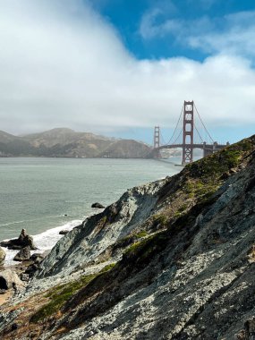 San Francisco 'daki Golden Gate köprüsünün kıyı manzarası. Amerikan simgesi. Uçurumlar ve çimenler resmi çerçeveliyor. Körfez suyunda dalgalar. Hafif sis.