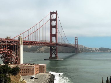 San Francisco 'daki Golden Gate köprüsünün kıyı manzarası. Amerikan simgesi. Uçurumlar ve çimenler resmi çerçeveliyor. Körfez suyunda dalgalar. Hafif sis.
