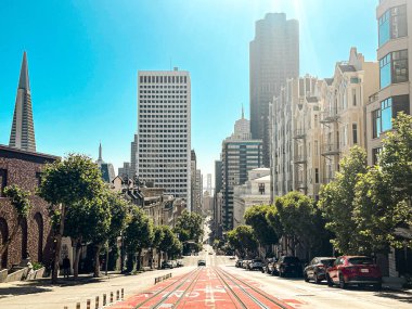 San Francisco 'daki tepenin tepesinden bak. Yolun ortasında, yolun aşağısına giden tramvay korkulukları var. Ufuktaki gökdelenler ve asma köprü.