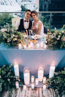 Genç bir çift düğün töreninde çiçekler ve mumlarla masada oturuyor.      