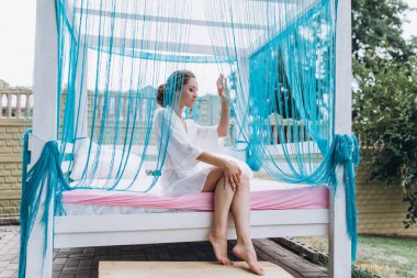 Beyaz bornozlu genç ve güzel bir kadın açık havada yatakta dinleniyor.       