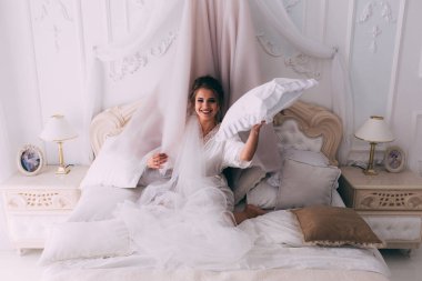 Güzel, mutlu, genç bir gelin yatak odasında gelinlikle poz veriyor. Gelinin özel sabahı..                     
