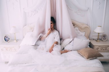 Güzel, mutlu, genç bir gelin yatak odasında gelinlikle poz veriyor. Gelinin özel sabahı..                     