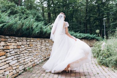 Beyaz gelinlikli güzel gelinin arka manzarası parktaki eski tuğla duvarın yanında yürüyor.