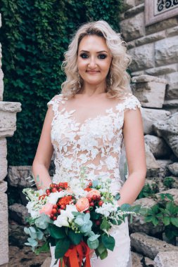 Beyaz gelinlik içinde yaşlı taş duvarın yanında poz veren mutlu, genç bir gelin.     