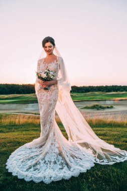 Şık elbiseli, elinde bir buket çiçekle gün batımında dışarıda poz veren güzel bir gelin.     