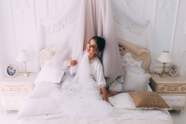 Güzel, mutlu, genç bir gelin yatak odasında gelinlikle poz veriyor. Gelinin özel sabahı..                     