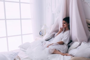 Güzel, mutlu, genç bir gelin yatak odasında gelinlikle poz veriyor. Gelinin özel sabahı..                     