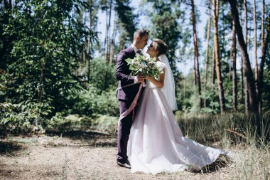 Mutlu genç bir çift ormanda yürüyor ve gülümsüyor. Buketli gelin        