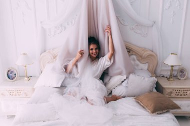 Güzel, mutlu, genç bir gelin yatak odasında gelinlikle poz veriyor. Gelinin özel sabahı..                     