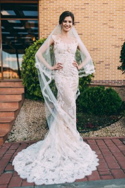 Güzel, mutlu, beyaz elbiseli, duvağı dışarıda poz veren genç gelin. 